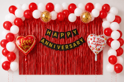 Red and white anniversary balloon decoration with red foil curtain backdrop, black gold Happy Anniversary banner, love you heart foil balloons and gold confetti balloons.