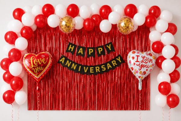 Red and white anniversary balloon decoration with red foil curtain backdrop, black gold Happy Anniversary banner, love you heart foil balloons and gold confetti balloons.