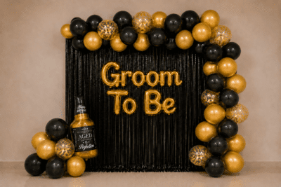 Groom to be party decoration with black and gold balloons, black foil curtain backdrop, golden Groom To Be foil banner, gold confetti balloons and a bottle foil balloon.