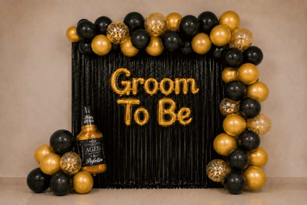 Groom to be party decoration with black and gold balloons, black foil curtain backdrop, golden Groom To Be foil banner, gold confetti balloons and a bottle foil balloon.