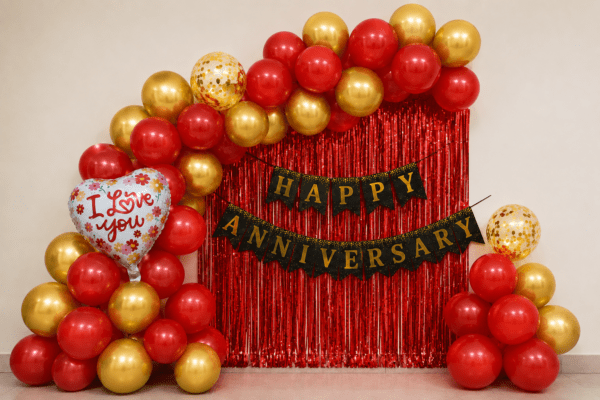 Red and gold Happy Anniversary balloon decoration with black gold banner, love you foil balloons and golden confetti balloons for anniversary party setup.