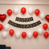 Red & White Happy Anniversary Balloon Decoration Red and white Happy Anniversary wall decoration with black and gold cardstock banner, 20 balloons and two red heart foil balloons for anniversary celebration.
