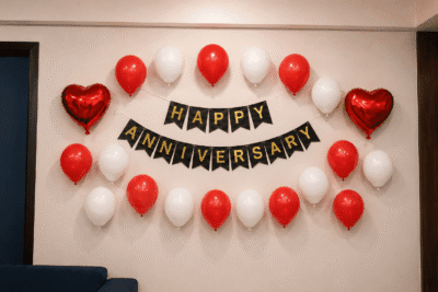 Red and white Happy Anniversary wall decoration with black and gold cardstock banner, 20 balloons and two red heart foil balloons for anniversary celebration.
